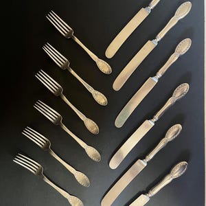 May include: Set of silver flatware, including forks and butter knives, arranged in a symmetrical pattern on a black background.
