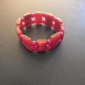 May include: A stretch bracelet composed of alternating rectangular orange-red stone beads and small round pink beads. The rectangular beads are arranged in a circular pattern, connected by the smaller pink beads, creating a continuous loop design.