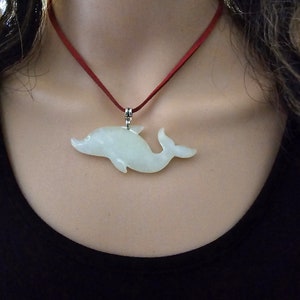 Puede incluir: Un colgante de delfín de jade blanco en un collar de cordón rojo. El delfín está tallado en un estilo realista y tiene un acabado liso y pulido.