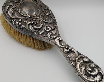 May include: An antique silver-coloured hairbrush with an ornate, swirling floral design. The brush head is oval, with natural-coloured bristles. The handle is integrated into the design, featuring the same detailed scrollwork.