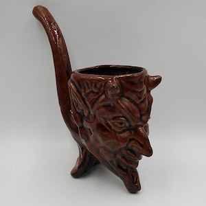 May include: A brown ceramic devil-faced pipe with a long, curved stem. The pipe features a detailed, textured face with horns and a wide grin. The bowl is dark and the overall design is a unique, novelty item.