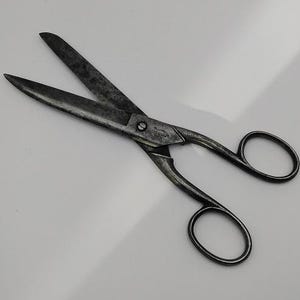 Puede incluir: Un par de tijeras antiguas con un acabado metálico oscuro. Las hojas están abiertas, mostrando un aspecto desgastado. Los mangos tienen bucles para los dedos. Las tijeras están sobre un fondo liso y claro.