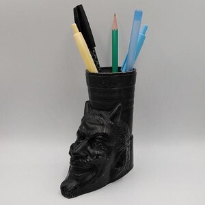 May include: Black desk organizer shaped like a devil's head with horns. The organizer holds a black pen, a yellow pen, a blue pen, and a green pencil.