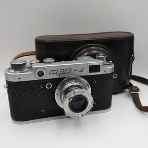 Puede incluir: Una cámara ФЭД-2 vintage en negro y plateado con una funda y correa de cuero marrón. La cámara tiene una lente y diales plateados, con el nombre del modelo impreso en la parte superior. La funda de cuero es marrón.