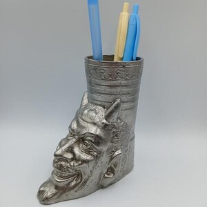 May include: A silver metal desk organizer shaped like a devil's head with horns. The organizer has three pens inside: one blue, one light blue, and one yellow.