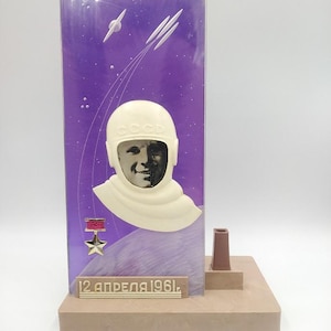 以下が含まれることがあります： 宇宙飛行士の肖像画が描かれた装飾彫刻。白いヘルメットをかぶり、紫色の背景に宇宙をテーマにしたグラフィックが描かれています。ベースには「12 АПРЕЛЯ 1961」と書かれています。