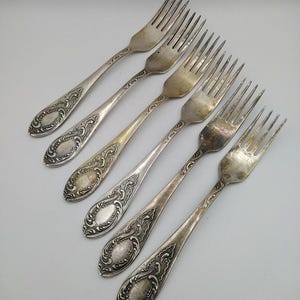 May include: Six antique silver forks arranged diagonally. Each fork features a decorative handle with an oval medallion and ornate scrollwork. The tines are slender and evenly spaced, showing signs of age and use.