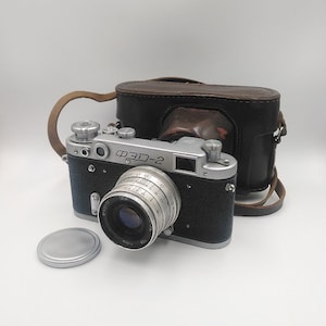 Puede incluir: Una cámara ФЭД-2 vintage con lente plateada y cuerpo negro, acompañada de una funda de cuero y tapa de lente. La cámara tiene un diseño clásico, con diales y perillas. La funda es marrón y negra.