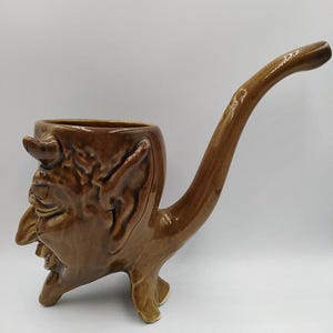 May include: A brown ceramic pipe shaped like a devil's head with a long, curved stem. The pipe has a detailed face with horns, ears, and a wide mouth.