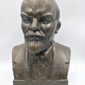 Könnte beinhalten: Eine bronzene Büste eines Mannes mit Glatze, markanter Stirn und gepflegtem Schnurrbart und Ziegenbart. Die Büste zeigt einen Mann in einem Anzug und Krawatte. Die Skulptur ist auf einem rechteckigen Sockel montiert.