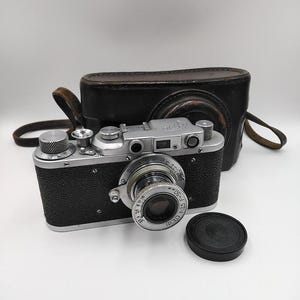 Könnte beinhalten: Eine Vintage-Kamera in Silber und Schwarz mit Objektiv, Zifferblättern und einer schwarzen Ledertasche. Die Kamera hat einen schwarzen Objektivdeckel und einen Lederriemen. Die Kamera befindet sich auf einer weißen Oberfläche.