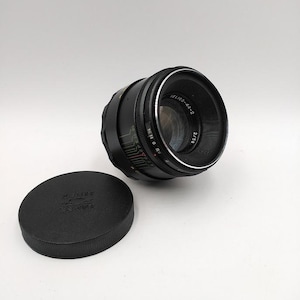 Op de afbeelding: Een zwarte cameralens met zilveren rand en de tekst "HELIOS-44-2" en "2/58" aan de zijkant. Een zwarte lensdop met het woord "ZENIT" staat naast de lens. De lens staat op een witte achtergrond.