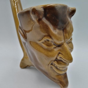 Puede incluir: Una taza de cerámica marrón con forma de cara de diablo con cuernos. La taza tiene una superficie texturizada y una expresión facial detallada.