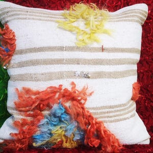 May include: White striped pillow with colorful fluffy embellishments. The pillow has three horizontal stripes of a light brown color. The embellishments are in shades of yellow, orange, and blue.