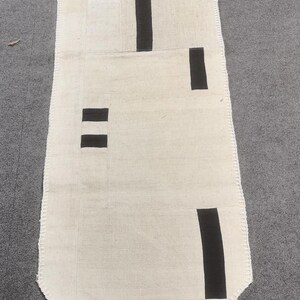 Puede incluir: Un camino de mesa blanco con diseños geométricos negros. El camino de mesa tiene un borde con flecos y una borla en el extremo.