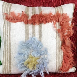 May include: A white pillow with brown stripes and a blue and yellow fluffy flower design. The pillow is decorated with orange fluffy trim.