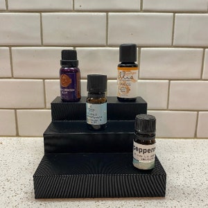 May include: Four small glass bottles with black caps and labels containing essential oils. The bottles are arranged on a black tiered stand with a textured surface. The bottles are labeled "Orange", "Eucalyptus", "Peppermint", and "Lavender"