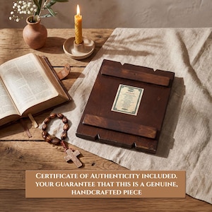 Pode incluir: &Iacute;cone religioso de madeira escura com certificado de autenticidade, sobre tecido bege. Um ros&aacute;rio com cruz, um livro aberto, uma vela acesa e um pequeno vaso com flores tamb&eacute;m s&atilde;o vis&iacute;veis. O texto diz: CERTIFICADO DE AUTENTICIDADE INCLU&Iacute;DO.