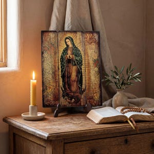 Pode incluir: Um &iacute;cone religioso de madeira retratando uma figura em um manto escuro com uma aur&eacute;ola, exibido em um suporte de madeira. Uma vela acesa em um suporte branco, um livro aberto e um vaso com vegeta&ccedil;&atilde;o est&atilde;o em uma mesa de madeira.