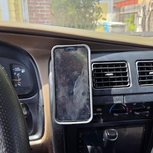 Puede incluir: Un smartphone en una funda plateada está montado en el salpicadero de un coche. El teléfono está en posición vertical, y el salpicadero tiene rejillas de ventilación, una radio y un volante. El interior es marrón y negro.