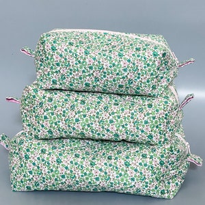 May include: Three rectangular cosmetic bags stacked. They feature a white fabric with a pattern of small green and pink flowers. Each bag has a zip closure and small pink fabric loops on the sides. The bags are made of quilted fabric.