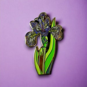 Peut inclure: Une épingle en émail dorée représentant une illustration détaillée d'une fleur d'iris violette avec des feuilles vertes. La fleur a un centre blanc et est entourée d'un contour doré.