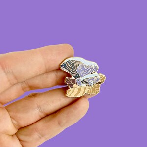 May include: A gold-toned enamel pin featuring a cluster of purple and blue mushrooms with brown and beige leaves. The mushrooms have a light blue and white gradient.