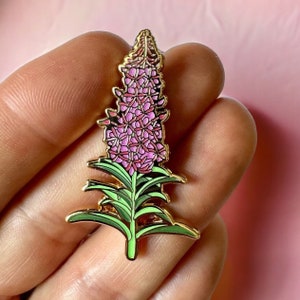 May include: A gold-tone enamel pin depicting a pink and green flower with intricate detail. The flower has a tall, slender stem and a cluster of pink blossoms.
