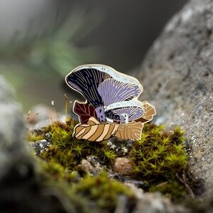 May include: A purple and gold enamel pin featuring a mushroom with a detailed design. The pin is set against a background of green moss.