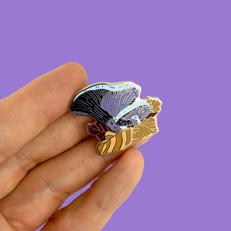 May include: A gold-tone enamel pin featuring a purple and blue mushroom with a light blue underside, nestled in brown and orange leaves.