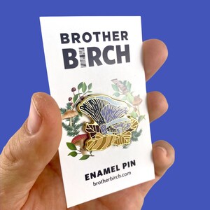 May include: A gold enamel pin featuring a purple and blue mushroom with a brown and gold leaf design. The pin is surrounded by a green and brown floral design. The text "BROTHER BIRCH" and "ENAMEL PIN" are printed on the card. The website address "brotherbirch.com" is printed below the text "ENAMEL PIN".