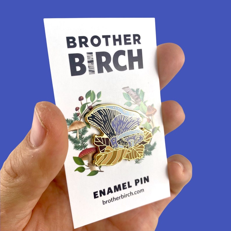 May include: A gold enamel pin featuring a purple and blue mushroom with a brown and gold leaf design. The pin is surrounded by a green and brown floral design. The text "BROTHER BIRCH" and "ENAMEL PIN" are printed on the card. The website address "brotherbirch.com" is printed below the text "ENAMEL PIN".