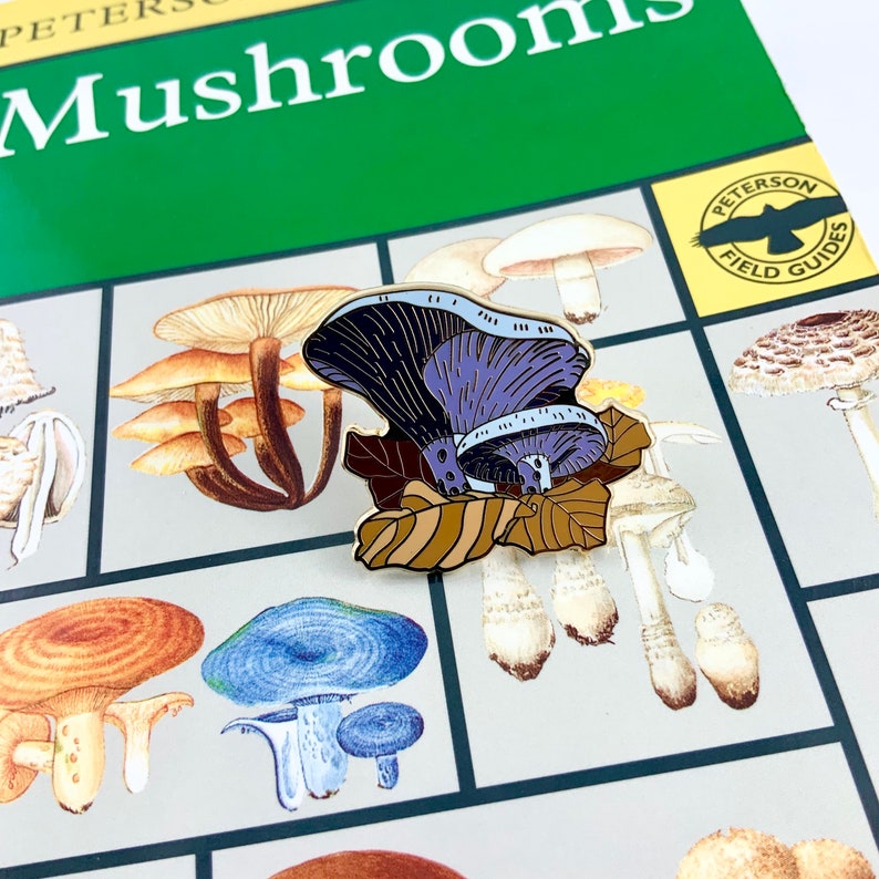 May include: A purple and black enamel mushroom pin with brown leaves. The pin is on top of a mushroom field guide book.