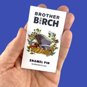 May include: A gold enamel pin featuring a purple and brown mushroom with green leaves and red berries. The pin says "Brother Birch" and "Enamel Pin" with the website "brotherbirch.com".