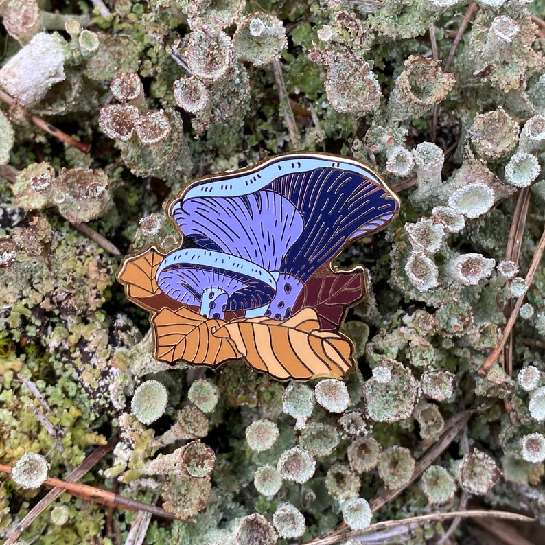 May include: A gold-tone enamel pin featuring a purple and blue mushroom with brown leaves. The pin is set against a background of green moss.