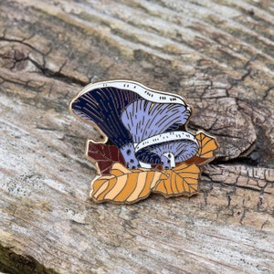 May include: A gold-tone enamel pin featuring a purple and blue mushroom with a white stem, surrounded by orange and brown leaves.
