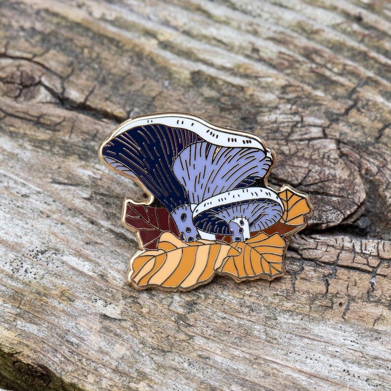May include: A gold-tone enamel pin featuring a purple and blue mushroom with a white stem, surrounded by orange and brown leaves.