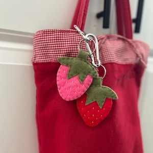 May include: A red tote bag with a gingham lining and a silver carabiner clip holding two felt strawberry keyrings. One strawberry is pink, the other is red, both with green tops.