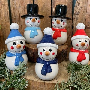 Könnte beinhalten: Sechs Keramik-Schneemannfiguren. Jeder Schneemann hat einen weißen Körper, eine Karottennase und ein lächelndes Gesicht. Sie tragen schwarze Zylinder oder blaue und rote Wintermützen sowie bunte Schals.