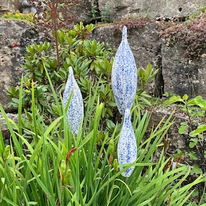 Könnte beinhalten: Drei blau-weiß gesprenkelte, tropfenförmige Gartenornamente stehen zwischen hohem, grünem Gras und Blättern. Die Ornamente sind hoch und schlank, mit einer spitzen Spitze. Eine Felswand und anderes Grün befinden sich im Hintergrund.