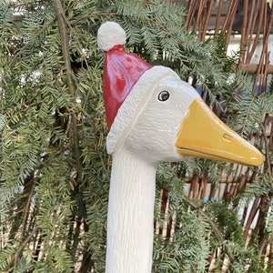 Könnte beinhalten: Eine weiße Keramik-Gans-Figur mit rotem Weihnachtsmütze und gelbem Schnabel. Die Gans steht vor einem Hintergrund aus grünem Laub, eine festliche Weihnachtsdekoration.