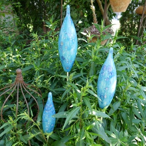 Könnte beinhalten: Vier blaue Keramik-Gartenstecker mit Tropfendesign werden inmitten von grünem Laub gezeigt. Die Stecker haben ein gesprenkeltes Muster in Blau- und Weißtönen. Ein rostiges dekoratives Metallelement ist ebenfalls sichtbar.
