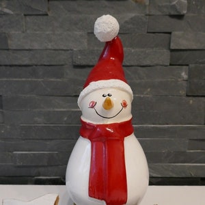 Könnte beinhalten: Eine weiße Keramik-Schneemann-Figur mit roter Weihnachtsmütze und Schal. Der Schneemann lächelt und hat eine Karottennase. Die Figur steht auf einem Holzsockel mit Tannenzapfen drumherum.