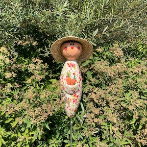 May include: A whimsical garden stake featuring a smiling face with a straw hat. The stake has a white body with a floral pattern and a pocket holding two orange carrots. The stake is set in a lush green garden.