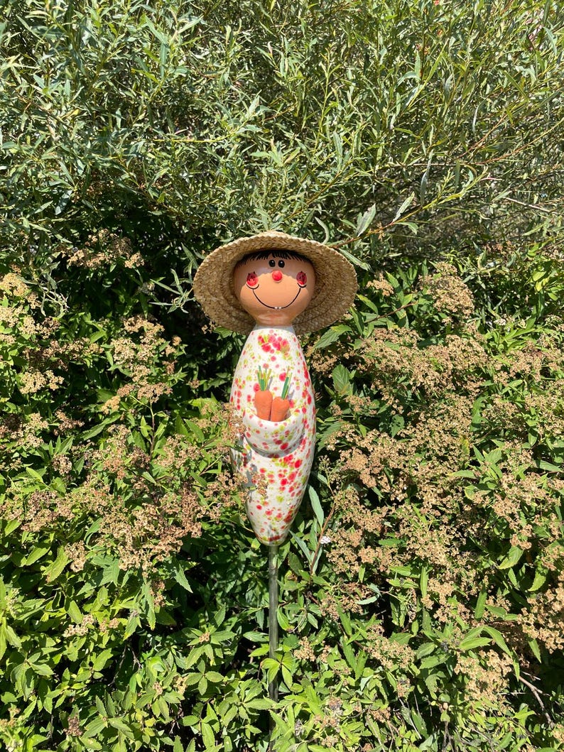 May include: A whimsical garden stake featuring a smiling face with a straw hat. The stake has a white body with a floral pattern and a pocket holding two orange carrots. The stake is set in a lush green garden.