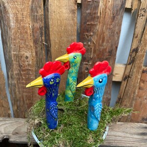 Könnte beinhalten: Drei Keramik-Hahnfiguren in Rot, Blau und Gelb bemalt. Die Hähne stehen in einem kleinen Metalltopf mit grünem Moos.