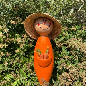 May include: A whimsical orange ceramic garden statue shaped like a person, with a straw hat and a smiling face. The statue holds two small carrots in a pocket. The statue is surrounded by green foliage.