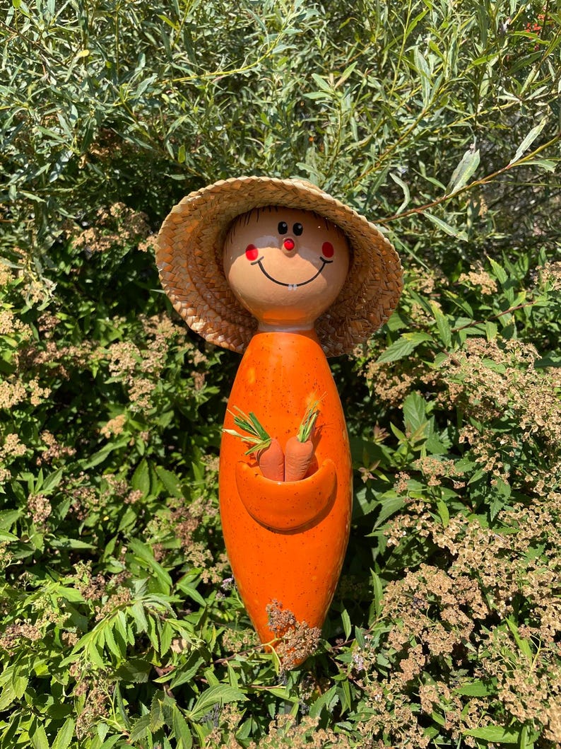 May include: A whimsical orange ceramic garden statue shaped like a person, with a straw hat and a smiling face. The statue holds two small carrots in a pocket. The statue is surrounded by green foliage.