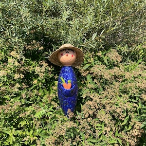 May include: A whimsical ceramic garden figurine of a smiling figure with a straw hat, blue polka-dotted body, and a pocket holding two orange carrots. The figurine is set amidst lush green foliage, creating a charming outdoor scene.
