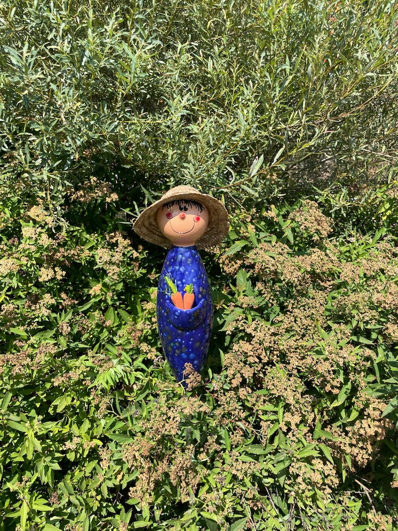 May include: A whimsical ceramic garden figurine of a smiling figure with a straw hat, blue polka-dotted body, and a pocket holding two orange carrots. The figurine is set amidst lush green foliage, creating a charming outdoor scene.